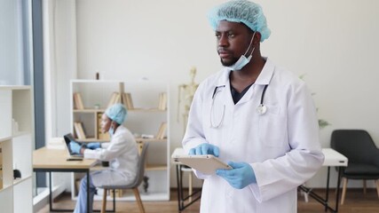 Male adult doctor wearing surgical attire and gloves reads medical information in clinic. Background nurse focuses on computer work, representing teamwork, medical professions - Powered by Adobe