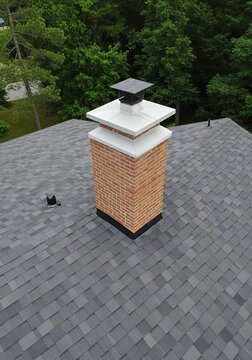 Aerial view of a modern brick chimney with a white crown and black cap on a grey asphalt shingle roof.
