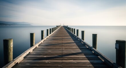Fototapeta premium Wooden pier stretches into still ocean under bright sky, distant hills