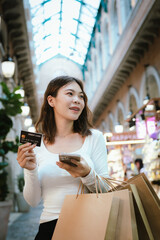 A happy young Asian woman smiles while holding shopping bags and a credit card, representing...