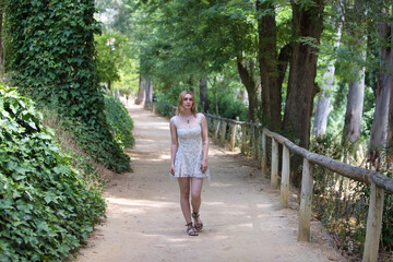 A young, beautiful blonde woman wearing a short floral print dress walks among trees along a path...