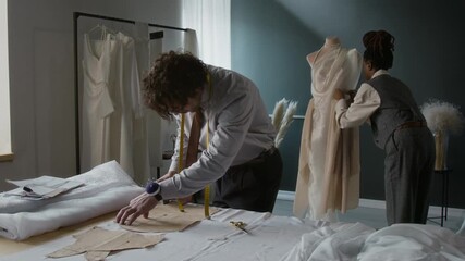 Young Caucasian man tracing sewing pattern, while his Black female colleague pinning fabrics onto mannequin