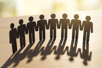 Paper-cut human figures holding hands in unity casting long shadows on wooden surface under warm...