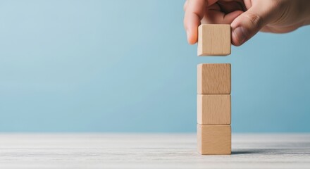 Hand placing a wooden block on a stack of blocks against a light blue backdrop
