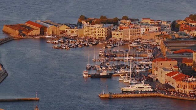 Sweeping aerial pan of Chania town, Crete, showcasing the Venetian Harbor, historic buildings, yachts, and golden sunlight during golden hour.