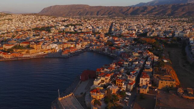 Aerial view of Chania, Crete, showcasing the Venetian Harbor, lighthouse, old town architecture, and mountains during golden hour with smooth motion.