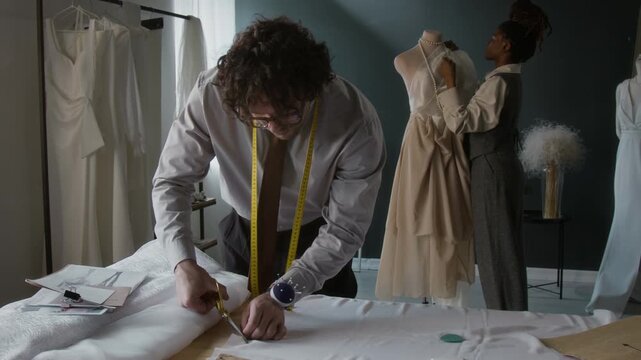Young male fashion designer cutting white fabric, while his female colleague using mannequin in background - Powered by Adobe