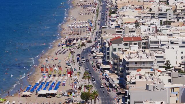 Aerial view of Rethymno, Crete, showing a sandy beach with umbrellas, turquoise sea, urban buildings, and rugged mountains in the background.