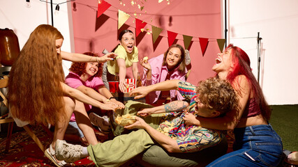 Group of young friends laughing and grabbing snacks at party with joyful chaotic fun. Concept of...