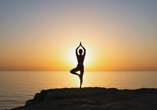 Woman in a warrior yoga pose on a cliff overlooking the ocean at sunrise for a wellness and spiritual concept - Powered by Adobe