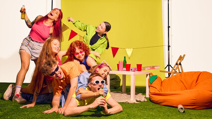 Pile of young friends stacked on grass with drinks and party smiles. Concept of carefree energy,...