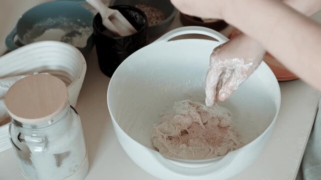 Close-up video of hands sprinkling salt into sourdough, then kneading the mixture in a white bowl on a clean kitchen counter.