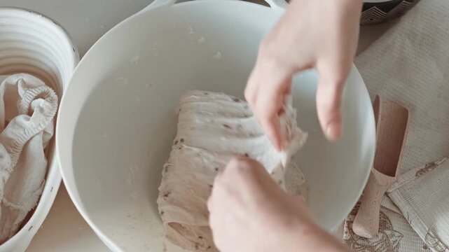 Close-up video of hands stretching and folding bread dough with seeds in a white bowl during sourdough bulk fermentation.