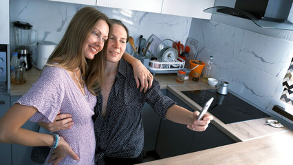 Two happy, joyful women take selfies in the kitchen. Lesbian lovers smile and have fun at home. Best friends take a photo together