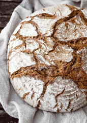 Golden and crusty, the round loaf rests on linen, its cracked surface full of character. Homemade traditions and countryside comfort. View from above.