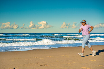 Beautiful mature woman training running on sandy beach in summertime. Physical activity on beach. Side view	