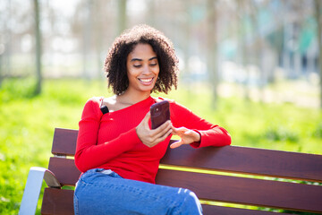 Smiling young woman sitting on a park bench outdoors, wearing a red sweater and using her smartphone on a sunny day