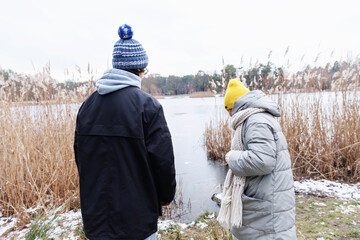 Teenage boy and his mother standing quietly by the edge of a frozen lake, dressed in warm winter...