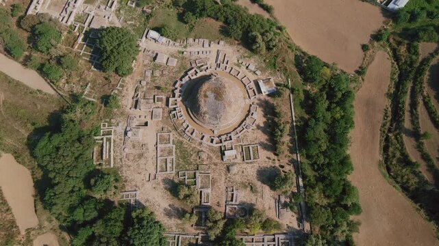 Aerial view of the ancient Dharmarajika Stupa ruins nestled among green trees and arid fields, a striking contrast of civilization and nature, Taxila, Punjab, Pakistan.