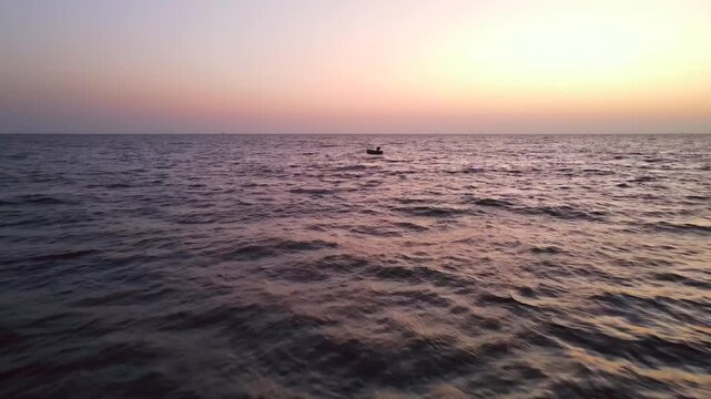 Aerial view of a lone boat sailing across the dark blue water, as the sun sets with a gradient of orange and pink tones, Karachi, Sindh, Pakistan.