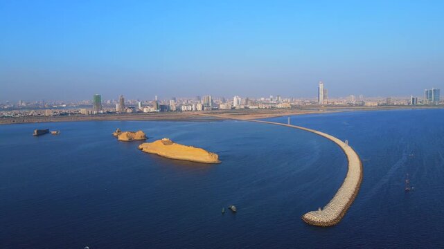 Aerial view of the contrast between the dark blue sea, the light brown islands, and the city skyline with a long breakwater, Karachi, Sindh, Pakistan.