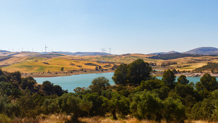 Naklejka premium Panoramic view of a rural landscape in Andalusia, with golden hills and a turquoise reservoir under a clear blue sky. The summer sun illuminates the landscape and a wind farm in the distance, adding a