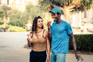 Happy multicultural couple walking outdoors eating ice cream cones together, smiling man and woman...