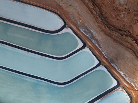 Aerial view of geometric evaporation ponds reflecting the sky, contrasting with the arid, red-brown earth, creating a surreal, patterned landscape, Location data omitted.