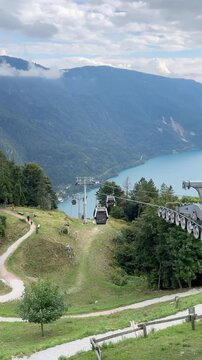 Time-lapse of cable car from Molva, Italy, ascending into the Alps in summer, with green meadows, lake, and mountain peaks viewed from above.