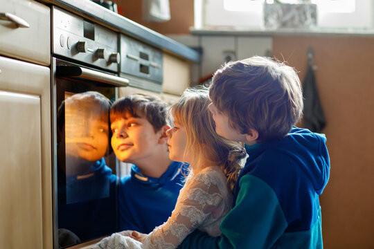 Little toddler girl and two kids boys baking pumpkin cake or cookies in kitchen. Three children, siblings, brothers and sister sitting near oven and waiting. On thanksgiving or christmas holiday.