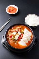 Korean food (Kimchi Jjigae), Kimchi soup with tofu, pork, scallion and chili in Korean stone bowl eating with cooked rice and kimchi on black background
