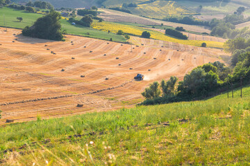 Closeup view of rolling golden hills dotted with hay bales and a tractor at work, bathed in warm...
