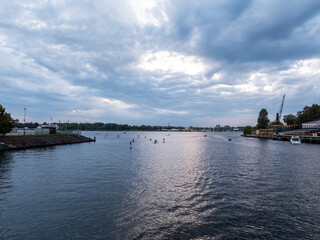 A wide river with paddleboarders, docked boats, and a crane on the right. The left shows a sloped...