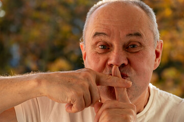 Senior male with playful expression makes shh gesture with finger to lips, signaling silence and...