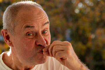 Senior male with playful expression makes shh gesture with finger to lips, signaling silence and...