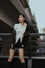 Stylish woman in a white shirt and black jacket standing confidently on a street railing.