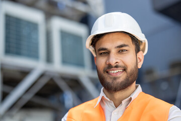 Smiling indian engineer wearing hardhat and safety vest in industrial setting