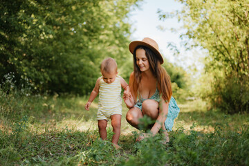 Fototapeta premium A smiling young mother in a summer dress and hat holds her little boy's hand as he takes his first steps in a grassy park on a sunny day.