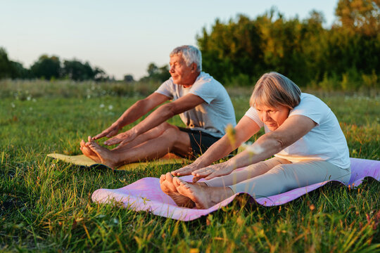 Mature couple in a seated forward bend pose, doing yoga in a grassy field during sunset.