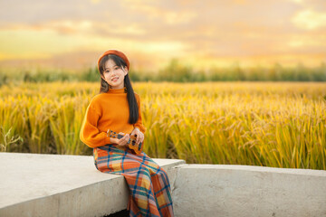 Teen girl in an orange sweater and plaid skirt playing ukulele in a golden field at sunset.