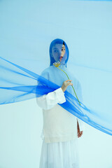 A young woman in a blue sweater holding a flower behind a sheer blue fabric.