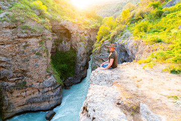 Traveler in hat sits on canyon edge with turquoise river, sunset golden light.