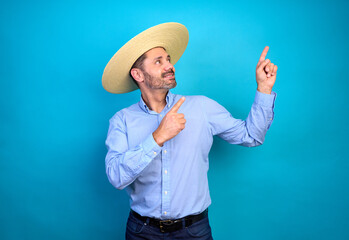Studio portrait of man wearing straw hat chupalla and light blue shirt pointing up with index fingers against a blue background, national holiday and patriotism celebration