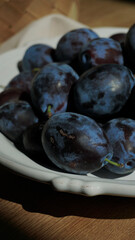 Close-up of fresh plums in a white ceramic bowl on a wooden surface. Summer harvest illuminated by...