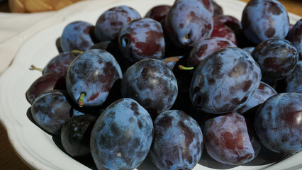 Close-up of fresh plums in a white ceramic bowl on a wooden surface. Summer harvest illuminated by...