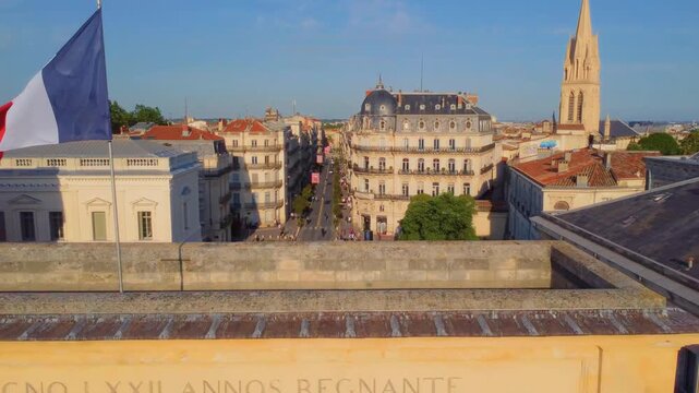 An aerial or elevated view captures the bustling Rue Foch in Montpellier, France, showcasing the iconic Porte du Peyrou (Arc de Triomphe) and the elegant Haussmannian style buildings lining the street