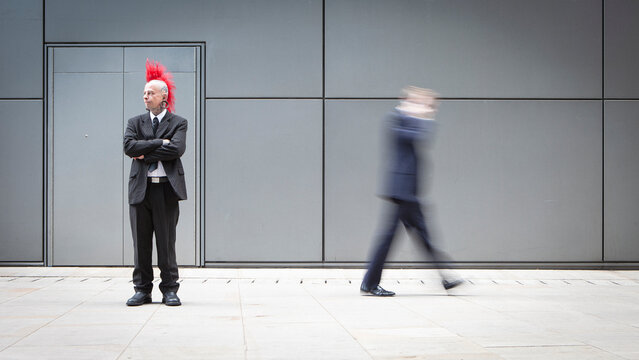Business Punk. A smartly dressed punk with a red mohawk stands still while a business man walks by in a blur. Full length portrait with copy space set in a contemporary business environment.