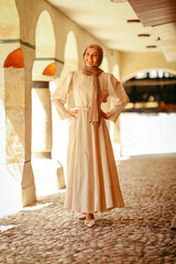 Naklejka premium Young woman in elegant modest dress and hijab posing confidently under historic stone arches, symbolizing culture, fashion and heritage.
