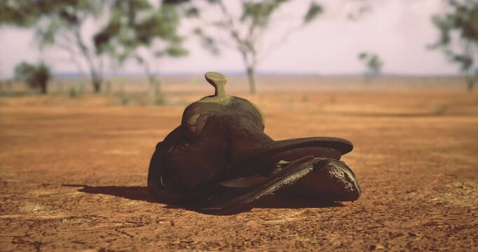 Amidst a barren landscape, a weathered cowboy saddle lies on the dry, cracked earth, surrounded by sparse vegetation and distant trees.