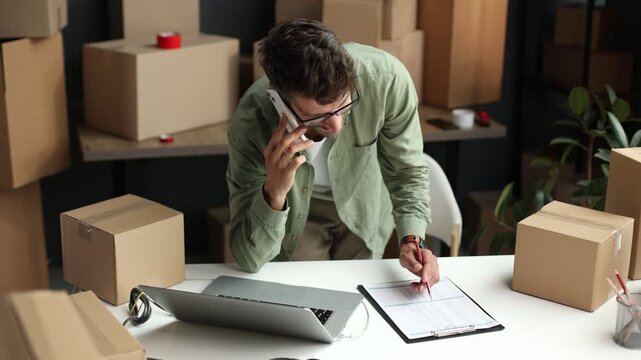 A man is talking on his cell phone while sitting at a desk with a stack of cardb